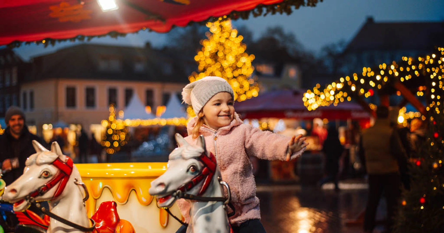 Kind mit Mütze und Jacke sitzt auf einem Karussellpferd auf dem Weihnachtsmarkt Bad Segeberg bei Dämmerung mit beleuchtetem Weihnachtsbaum im Hintergrund
