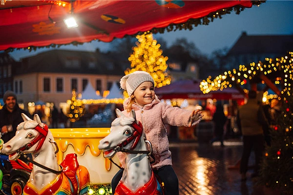 Kind mit Mütze und Jacke sitzt auf einem Karussellpferd auf dem Weihnachtsmarkt Bad Segeberg bei Dämmerung mit beleuchtetem Weihnachtsbaum im Hintergrund