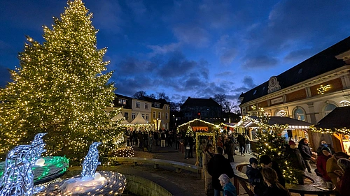 Weihnachtsmarkt bei Abenddämmerung mit großem, beleuchtetem Weihnachtsbaum und Menschen vor festlich geschmückten Buden