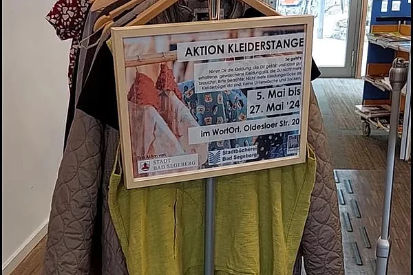 Kleiderstange mit mehreren Kleidungsstücken und einem Schild mit der Aufschrift 'AKTION KLEIDERSTANGE' in einem hellen Raum mit Glastür im Hintergrund