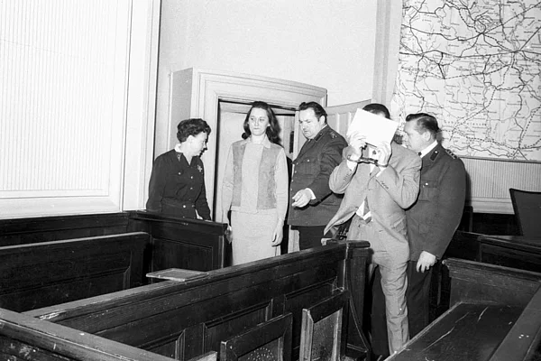 Fünf Personen in einem Gerichtssaal, darunter zwei in Uniform, eine Person hält ein Dokument vor das Gesicht