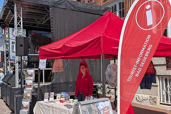 Roter Pavillon mit Infostand und Werbetafel vor rotem Backsteingebäude bei sonnigem Himmel