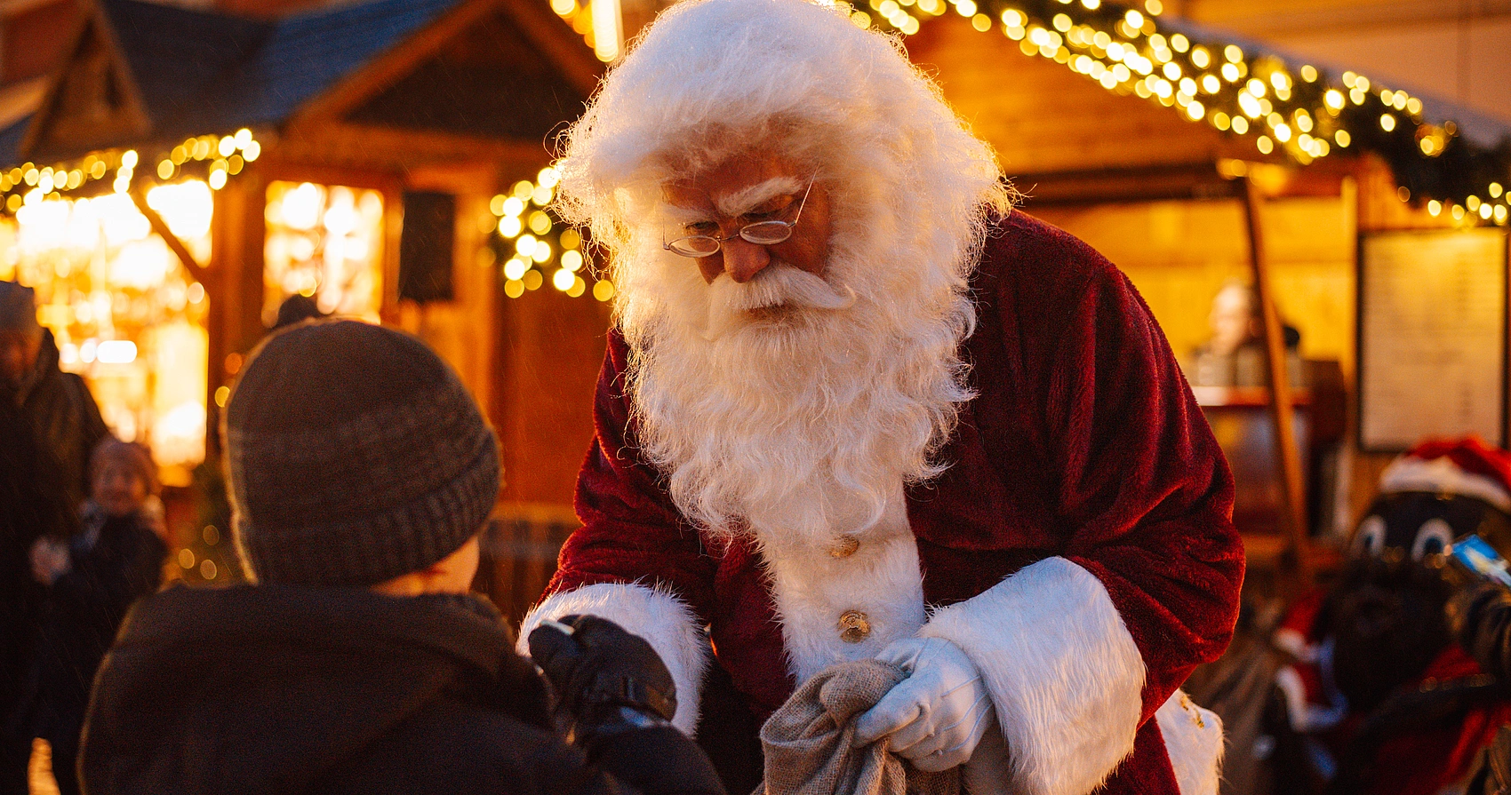 Weihnachtsmann mit weißem Bart und rotem Mantel übergibt einem Kind mit grauer Mütze auf einem Weihnachtsmarkt etwas aus einem Sack