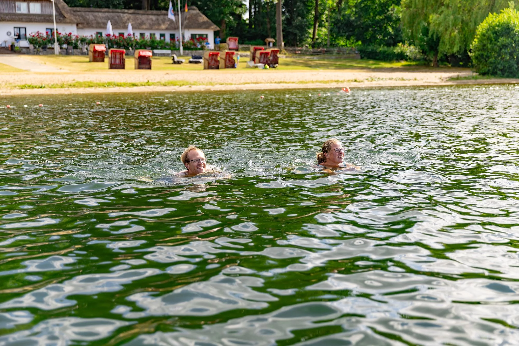 Zwei Personen schwimmen in einem See vor einem Ufer mit Sandstrand, Strandkörben und einem Gebäude im Hintergrund