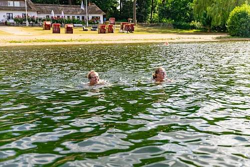 Zwei Personen schwimmen in einem See vor einem Ufer mit Sandstrand, Strandkörben und einem Gebäude im Hintergrund