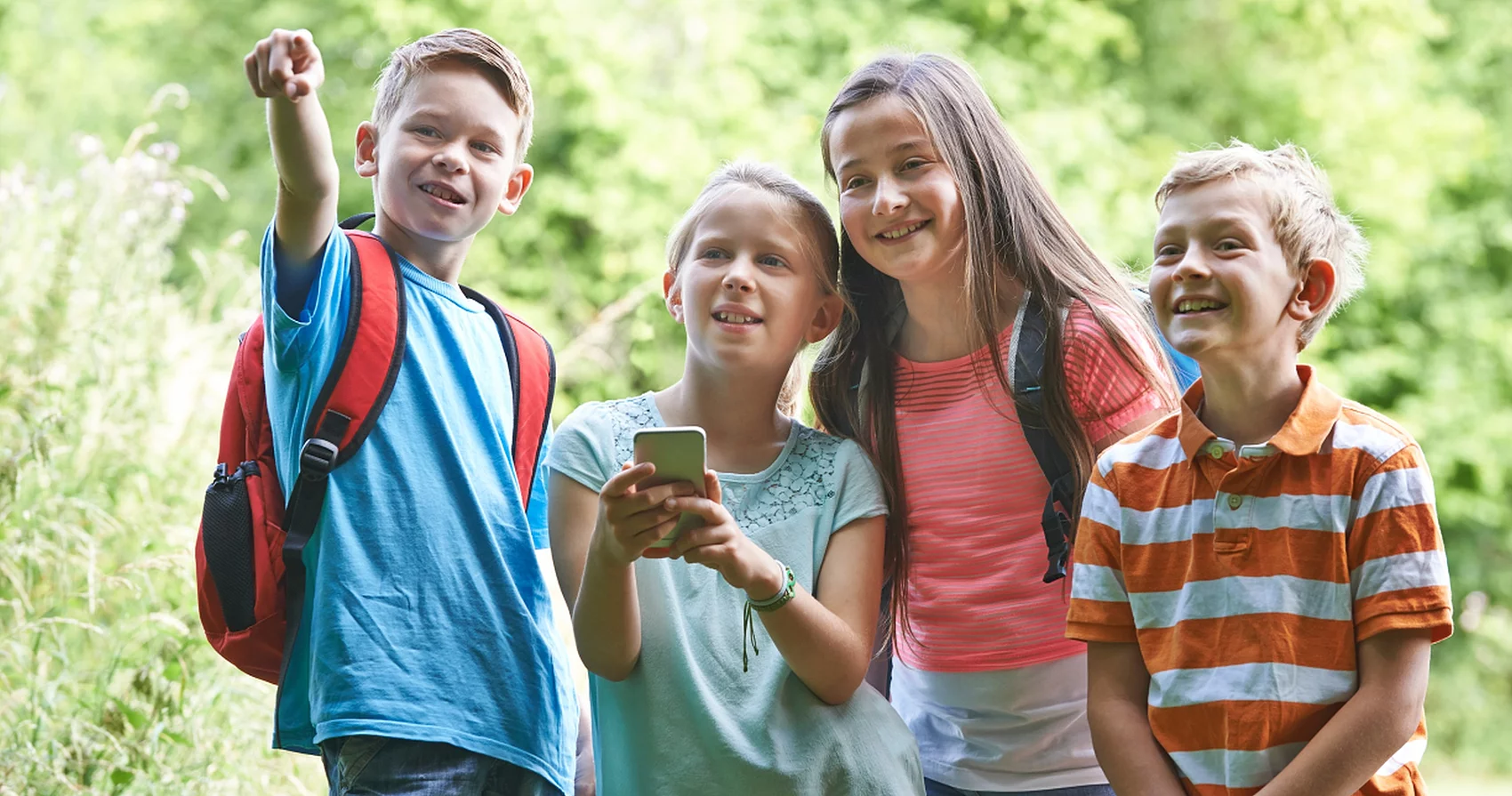Vier Kinder stehen im Freien, eines zeigt mit ausgestrecktem Arm nach vorne, ein anderes hält ein Smartphone, alle tragen sommerliche Kleidung und Rucksäcke