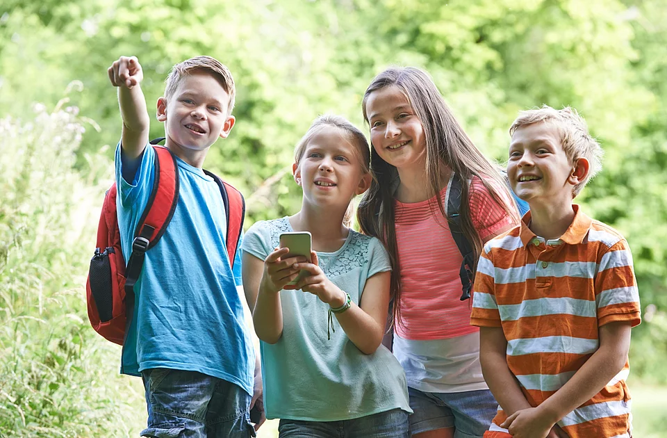 Vier Kinder stehen im Freien, eines zeigt mit ausgestrecktem Arm nach vorne, ein anderes hält ein Smartphone, alle tragen sommerliche Kleidung und Rucksäcke
