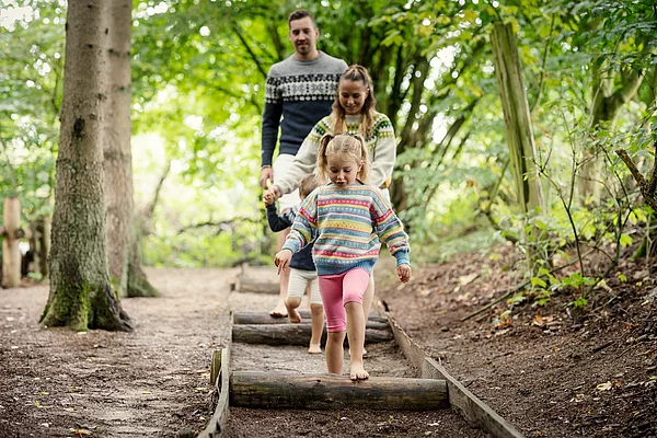 Vier barfüßige Personen in bunter Kleidung gehen hintereinander auf einem Barfuß-Waldweg über Baumstämme