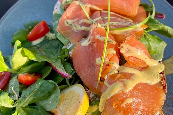 Offenes Brot mit Lachs, Salat, Kirschtomaten, Zitronenscheiben und Sauce auf grauem Teller