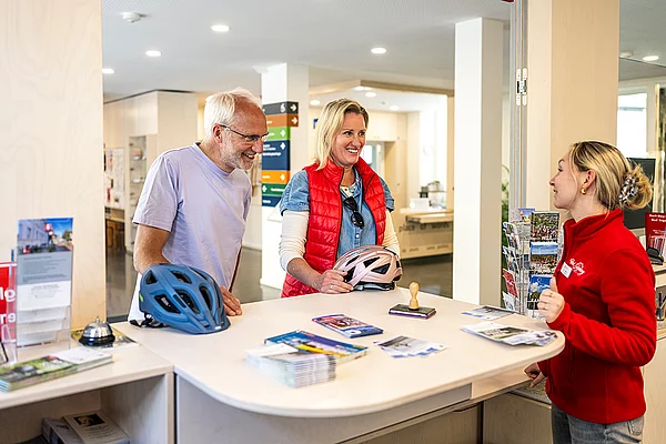 Drei Personen an einem Informationsschalter mit Fahrradhelmen auf der Theke und Broschüren