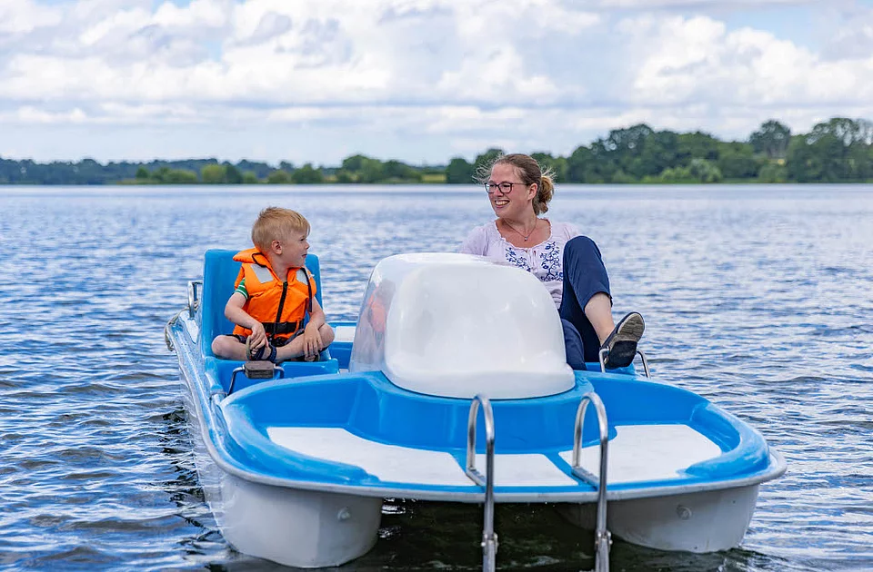 Kind mit orangefarbener Schwimmweste und Frau sitzen auf einem blauen Tretboot auf dem Großen Segeberger See