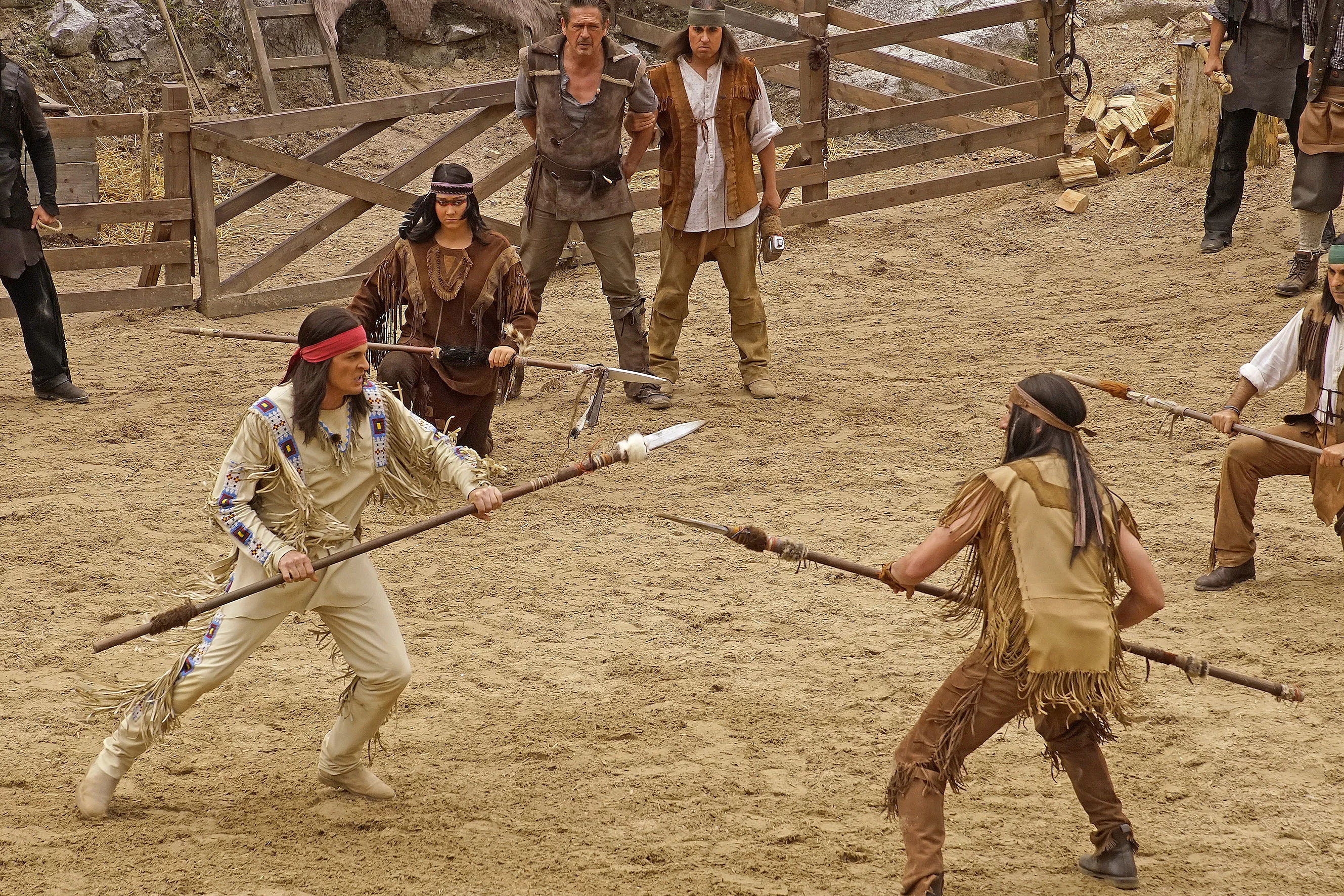 Zwei Männer in Indianer-Kostümen kämpfen mit Speeren auf einem sandigen Platz vor einem Holzzaun, umgeben von weiteren Personen in ähnlicher Kleidung