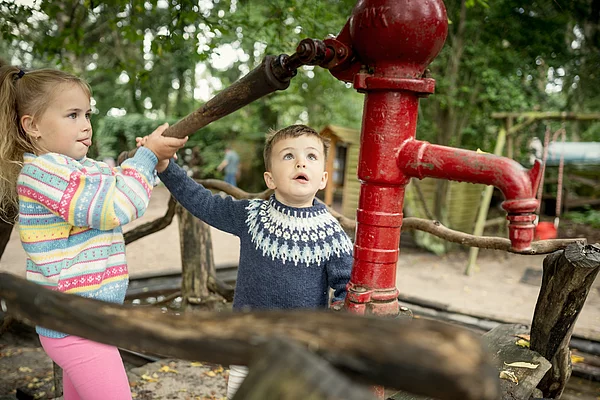Zwei Kinder betätigen gemeinsam eine rote Handpumpe im Freien