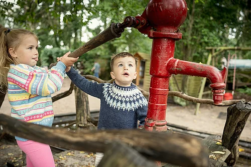 Zwei Kinder betätigen gemeinsam eine rote Handpumpe im Freien