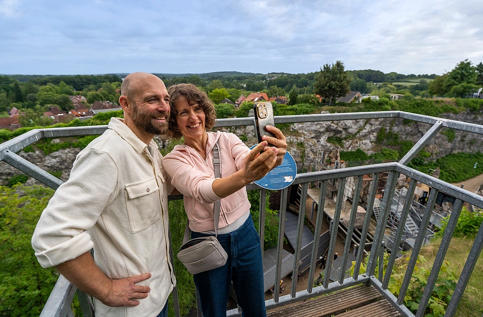 Zwei lächelnde Personen machen auf einer Aussichtsplattform mit Geländer ein Selfie vor einer grünen Landschaft und einem Dorf im Hintergrund