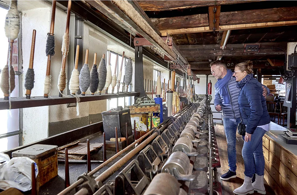 Zwei Personen betrachten in der Wollspinnerei Blunck in Bad Segeberg eine lange Spinnmaschine mit mehreren Spulen und Garnwicklungen an Holzstäben.