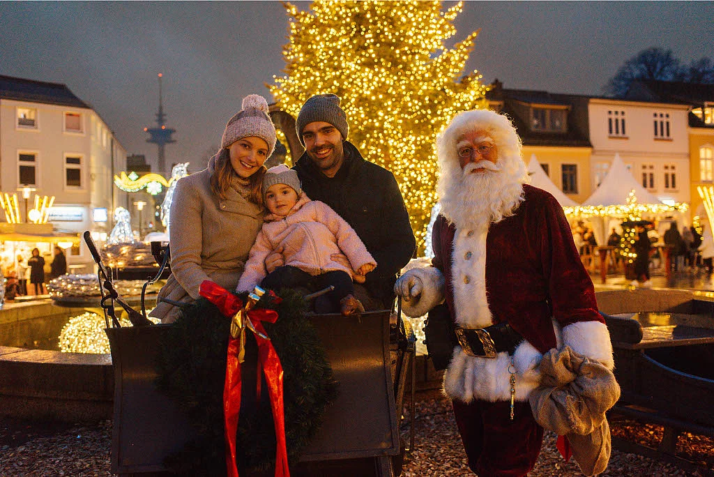 Familie mit Kind sitzt in einer festlich geschmückten Kutsche vor einem beleuchteten Weihnachtsbaum, daneben steht eine Person in Weihnachtsmannkostüm auf dem Weihnachtsmarkt Bad Segeberg