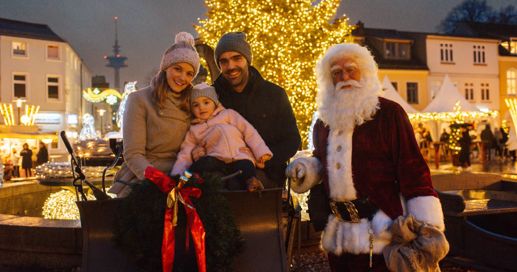 Familie mit Kind sitzt in einer festlich geschmückten Kutsche vor einem beleuchteten Weihnachtsbaum, daneben steht eine Person in Weihnachtsmannkostüm auf dem Weihnachtsmarkt Bad Segeberg