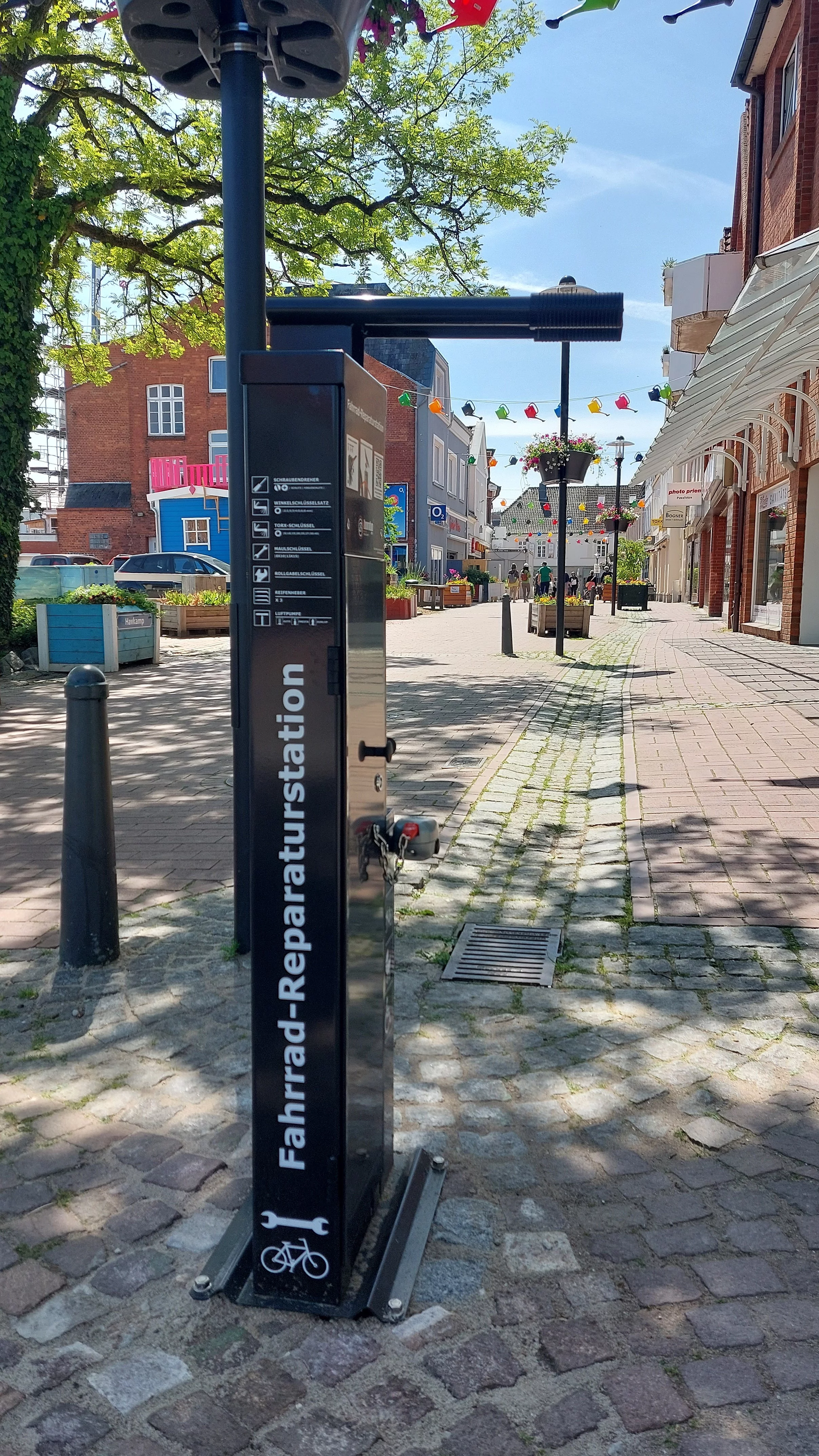 Fahrrad-Reparaturstation mit Werkzeugen an einer gepflasterten Straße in einer belebten Einkaufsstraße mit Geschäften und Bäumen