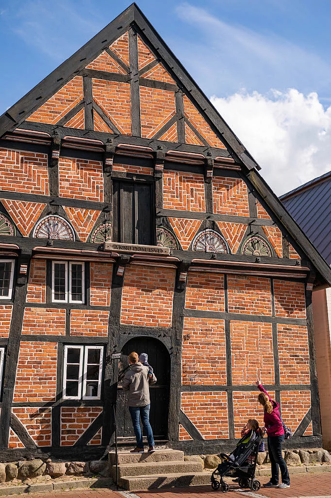 Fachwerkhaus mit rotem Ziegelmauerwerk und schwarzem Holzfachwerk, darüber eine verschlossene Tür im Giebel, davor eine Person mit Kind auf dem Arm an der Eingangstür und eine weitere Person mit Kinderwagen, die nach oben zeigt