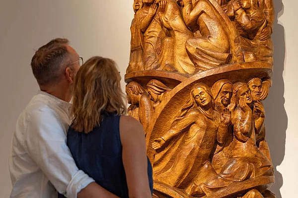 Zwei Personen betrachten eine hohe, vertikal gegliederte Holzskulptur mit mehreren reliefartigen, menschlichen Figuren in verschiedenen Posen in der Kunsthalle Otto Flath Bad Segeberg