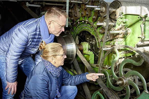 Zwei Personen in blauen Jacken und Schals betrachten und zeigen auf ein großes grünes historisches Spinnmaschinenrad in der Wollspinnerei Blunck Bad Segeberg