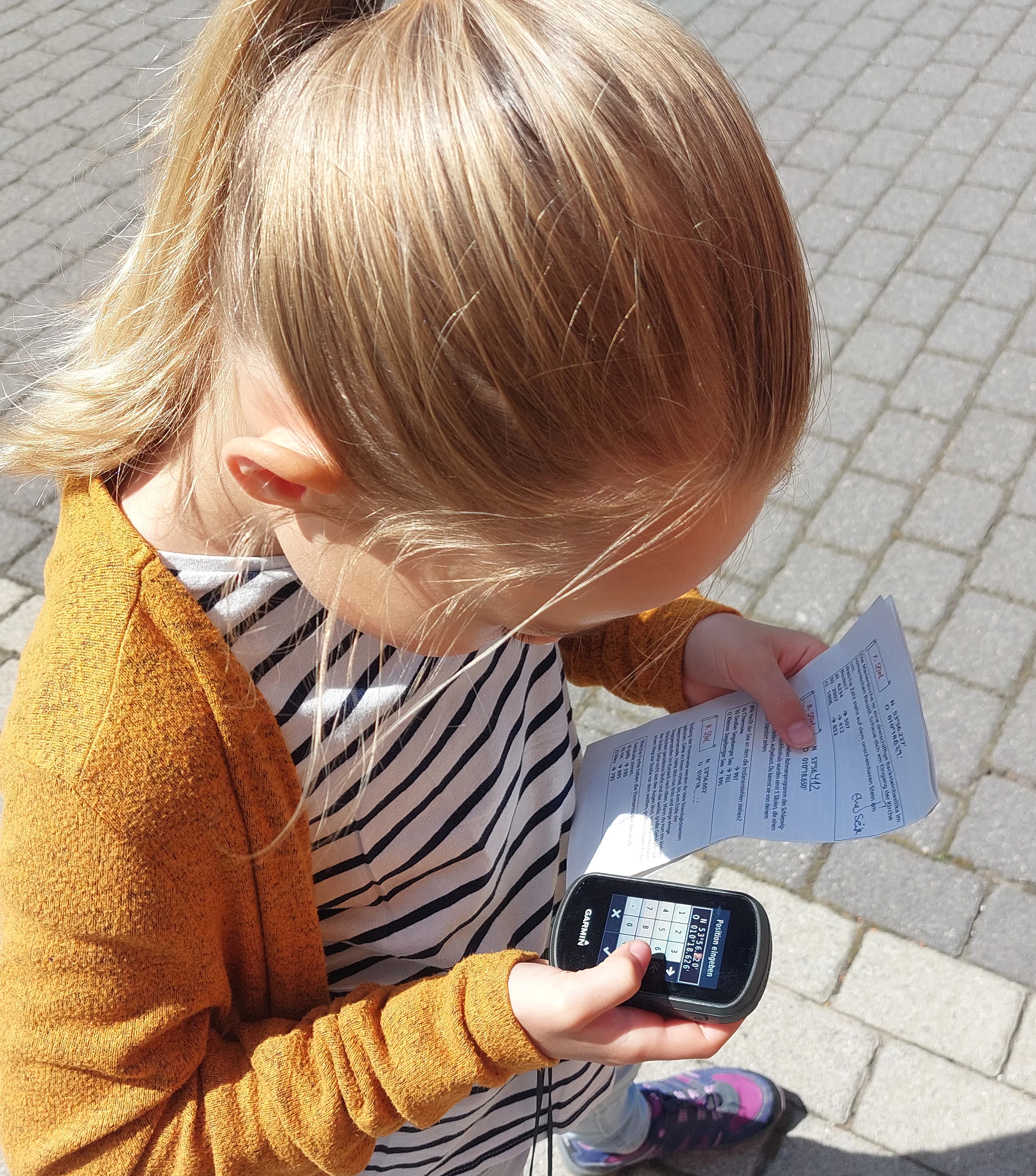 Kind mit blondem Pferdeschwanz steht auf gepflastertem Boden und hält ein GPS-Gerät sowie einen Zettel in den Händen