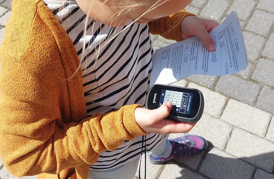 Kind mit blondem Pferdeschwanz steht auf gepflastertem Boden und hält ein GPS-Gerät sowie einen Zettel in den Händen