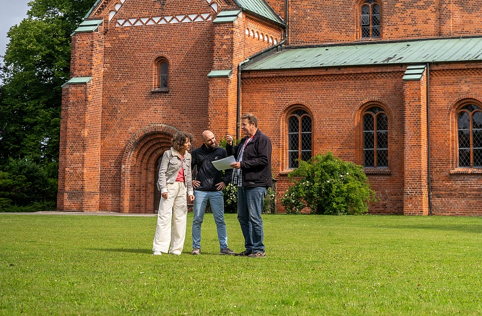 Drei Personen stehen auf einer grüen Wiese vor einer Kirche