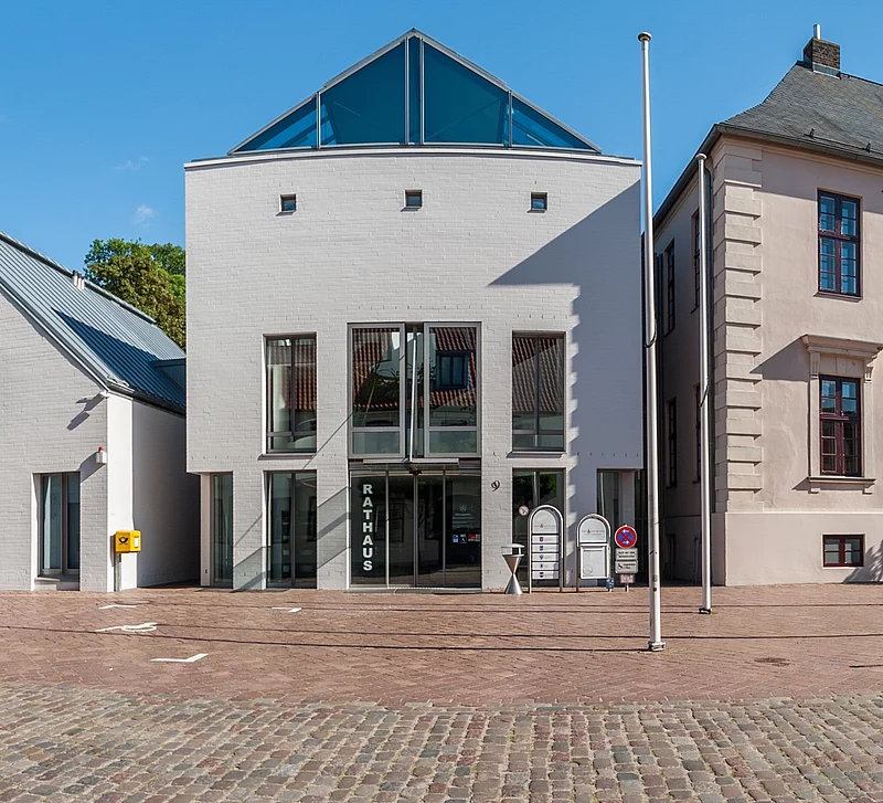 Straßenszene mit mehreren historischen und modernen Gebäuden, darunter das Rathaus von Bad Segeberg mit weißer Fassade und Glasfront, bei blauem Himmel