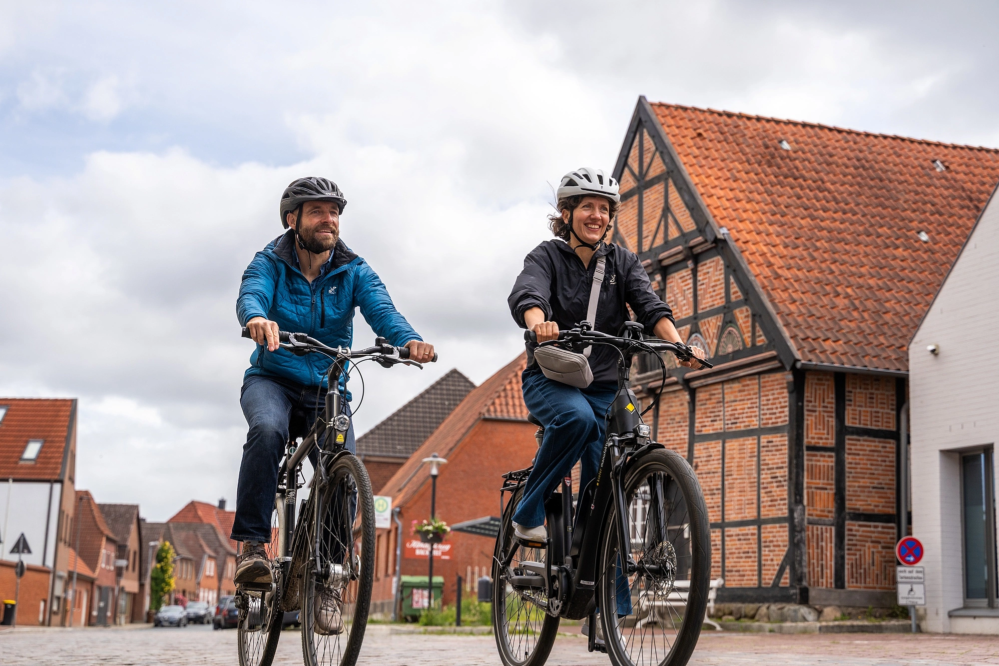 Zwei Personen mit Fahrradhelmen fahren auf E-Bikes durch eine Straße mit Fachwerkhäusern im Hintergrund