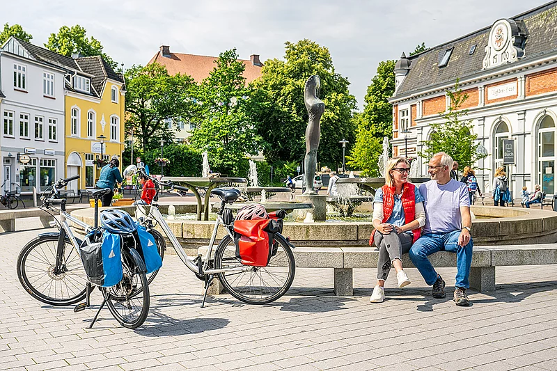 Zwei Fahrräder mit Packtaschen stehen vor einem runden Brunnen auf dem Marktplatz Bad Segeberg, daneben sitzt ein Paar auf der Brunnenmauer, umgeben von historischen Gebäuden und Bäumen.