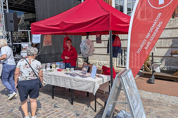 Roter Pavillon mit Infostand und rotem Fahnenbanner auf gepflastertem Platz vor Backsteingebäude, mehrere Personen stehen davor