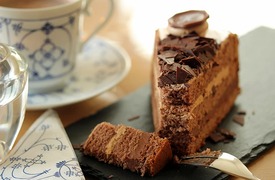 Ein Stück Schokoladenkuchen mit Schokoladenglasur und Schokoraspeln auf einer schwarzen Steinplatte neben einer Gabel und einer Tasse mit blau-weißem Muster.