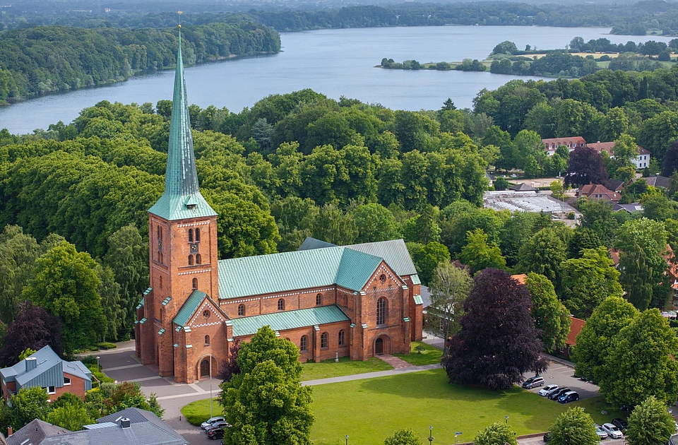 Luftaufnahme einer roten Backsteinkirche mit grünem Dach und hohem Turm umgeben von Bäumen und einem See im Hintergrund
