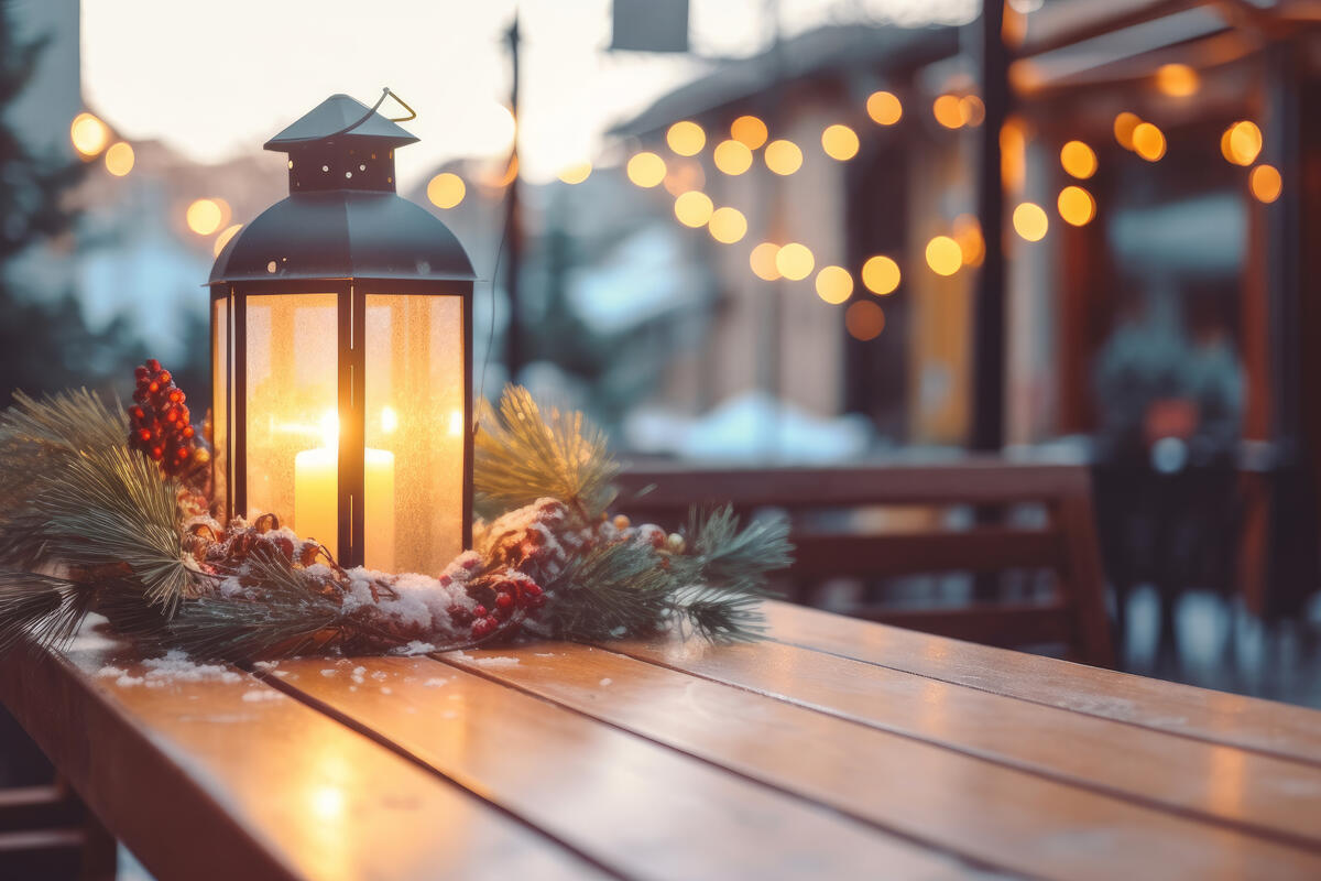 Eine Laterne mit einer leuchtenden Kerze steht auf einem Holztisch. Die Laterne ist mit Tannen geschmückt. Im Hintergrund hängen Lichterketten. 
