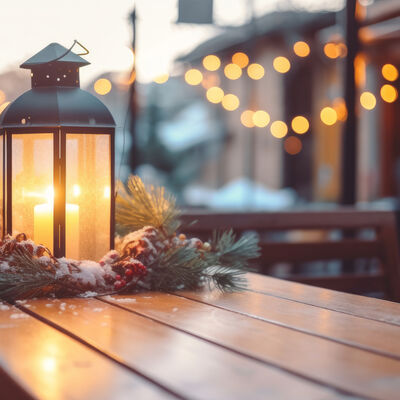 Eine Laterne mit einer leuchtenden Kerze steht auf einem Holztisch. Die Laterne ist mit Tannen geschmückt. Im Hintergrund hängen Lichterketten. 
