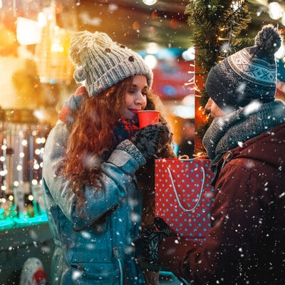 Bild vergrößern: Zwei Personen mit Winterjacke und Mtze stehen vor einem Weihnachtsmarkt-stand und trinken aus roten Bechern.