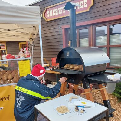 Bild vergrößern: Ein Mann mit roter Mütze hoclt vor einem mobilen Holzbackofen und schaut in den Ofen.