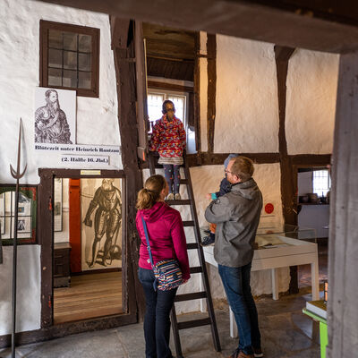 Bild vergrößern: Zwei Erwachsene stehen in einem historisch anmutendem Raum. Ein Mädchen steigt eine Holzleiter hoch.