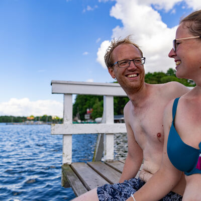 Bild vergrößern: Ein Mann und eine Frau sitzen in Badekleidung auf einem Steg und schauen sich lachend an.