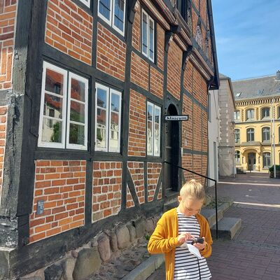 Bild vergrößern: Ein blondes Mdchen mit einer gelben Strickjacke steht vor einem alten Fachwerkhaus. Das Mdchen hlt ein kleines schwarzes Gert in der Hand.