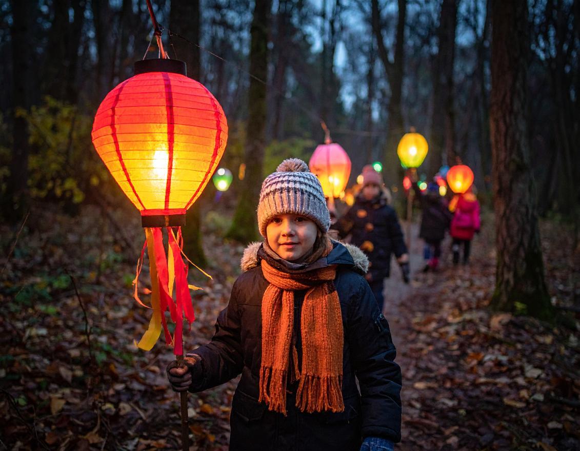 Ein Kind mit Wollmütze, Schal und Winterjacke hält eine orange leuchtende Laterne in der Hand. Im Hintergrund sind Wald in der Dämmerung und weitere Kinder mit Laternen zu sehen.