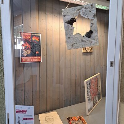 Bild vergrößern: Schaufenster mit Kunstwerken: Hängendes, zerrissenes Papier mit schwarzen Objekten, zwei gemalte Porträts auf dem Boden, ein Plakat mit Flammen und Text 'Damal Bücher - heute Menschen: Wenn Regierungen hassen, brennen Rechte', ein Flyer mit Text 'Kultur im Leerstand'.