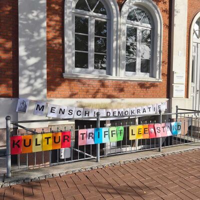 Bild vergrößern: Hinweisschild mit den Worten 'MENSCH! DEMOKRATIE' und bunte Buchstaben auf Papier mit dem Text 'KULTUR TRIFFT LEEESTAND' vor einem Kellereingang mit roten Backsteinwänden und zwei weißen Fensterbögen.