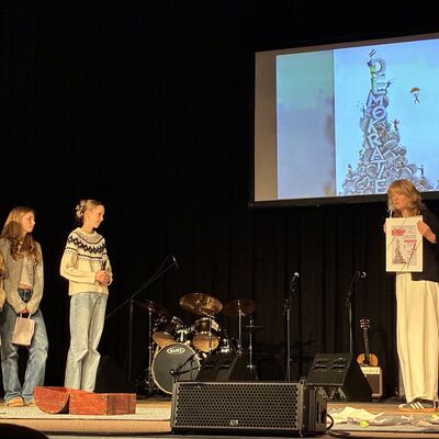 Bild vergrößern: Drei Jugendliche stehen auf der Bühne des Kleinen Theaters in Wahlstedt, eine erwachsene Frau hält ein Plakat mit einer Zeichnung und dem Wort 'Demokratie' vor einer Projektion mit demselben Motiv.