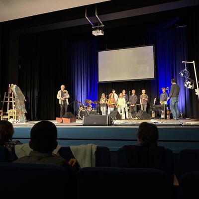 Bild vergrößern: Bühne im Kleinen Theater Wahlstedt mit mehreren Personen, die vor einem blauen Vorhang stehen, Musikinstrumente und Staffeleien mit Kunstwerken sichtbar.