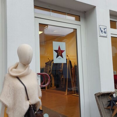 Schaufenster eines Bekleidungsgeschäfts mit Tür, darauf ein weißer Stern mit roter Zahl '12', davor Schaufensterpuppe mit cremefarbenem Poncho und brauner Tasche, daneben Korb mit getrockneten Blumen.