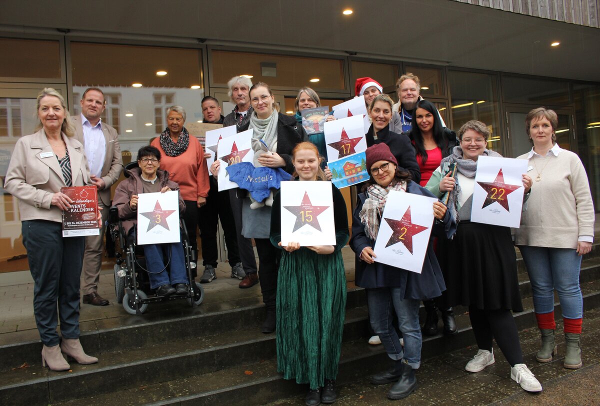 Eine Gruppe von Menschen steht auf einer Treppe vor einem Gebäude. Einige halten weiße Schilder mit roten Sternen und Zahlen, die an einen Adventskalender erinnern. Die Zahlen auf den Schildern sind unter anderem 2, 3, 15, 17 und 22. Eine Person trägt eine rote Weihnachtsmütze. Die Kleidung der Personen ist unterschiedlich, von formell bis leger. Im Hintergrund sind Glastüren des Gebäudes zu sehen.