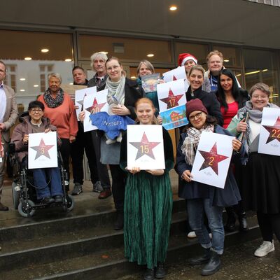 Bild vergrößern: Eine Gruppe von Menschen steht auf einer Treppe vor einem Gebude. Einige halten weie Schilder mit roten Sternen und Zahlen, die an einen Adventskalender erinnern. Die Zahlen auf den Schildern sind unter anderem 2, 3, 15, 17 und 22. Eine Person trgt eine rote Weihnachtsmtze. Die Kleidung der Personen ist unterschiedlich, von formell bis leger. Im Hintergrund sind Glastren des Gebudes zu sehen.